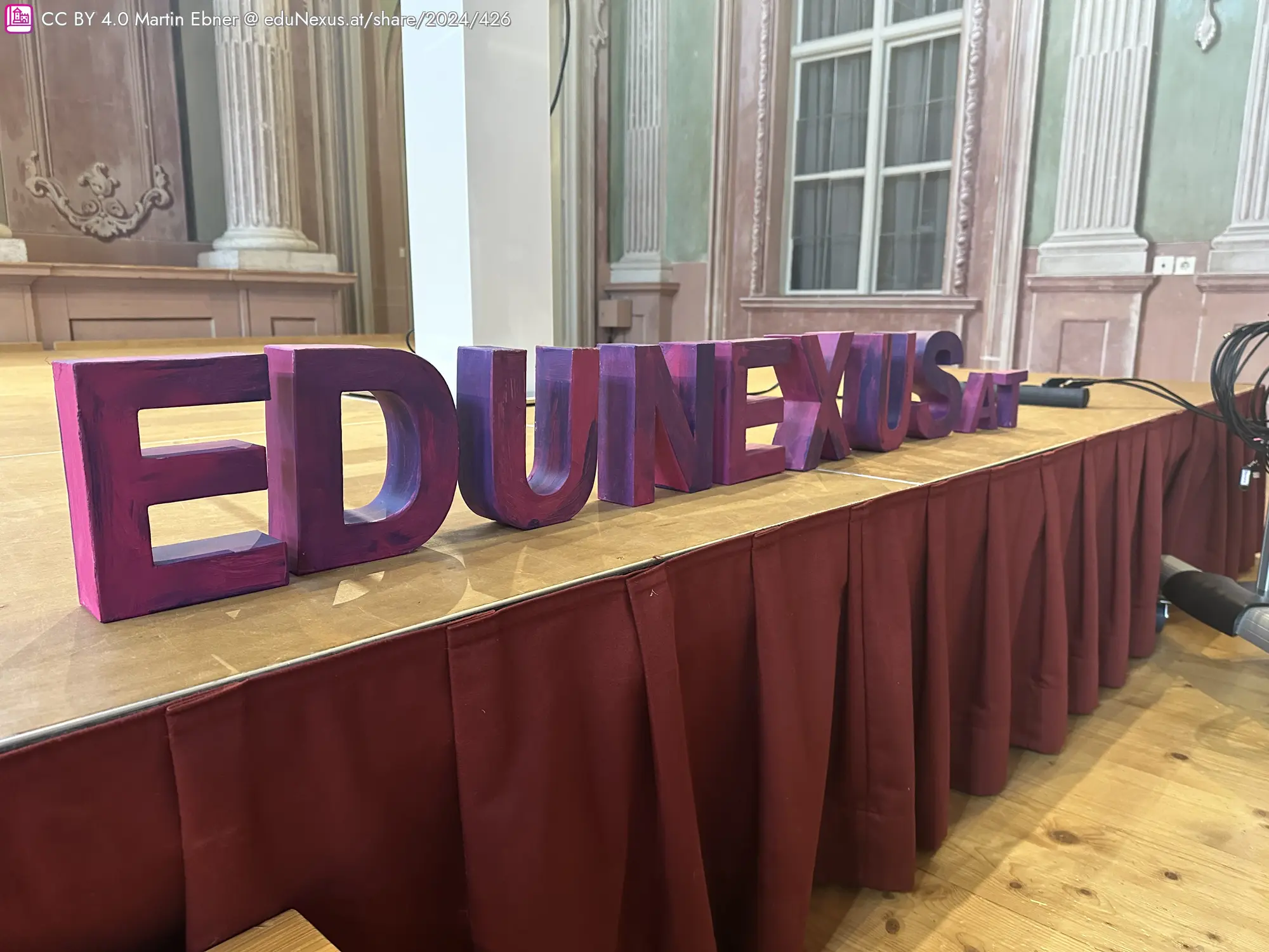 Bunte 3D-Buchstaben „EDUNEXUS“ auf einem Tisch mit rotem Tuch, im Hintergrund eine elegante Innenarchitektur.