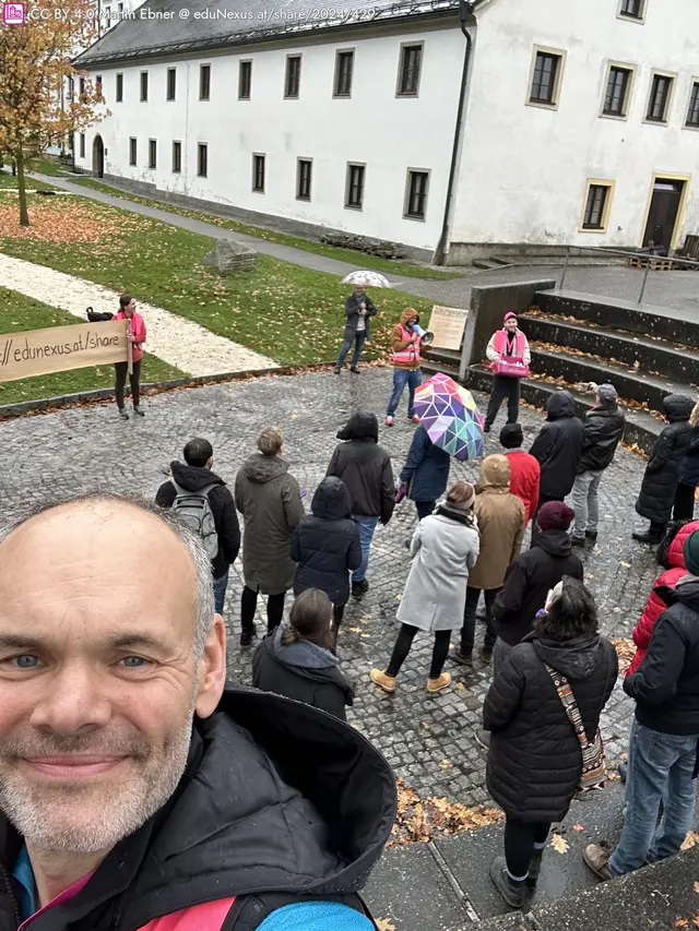 Gruppe von Menschen in Jacken und Regenschirmen, die in einem Innenhof stehen. Im Vordergrund ein Mann mit Glatze und Bart. Im Hintergrund ein Gebäude mit weißen Wänden und Fenstern. Herbstlaub auf dem Boden.