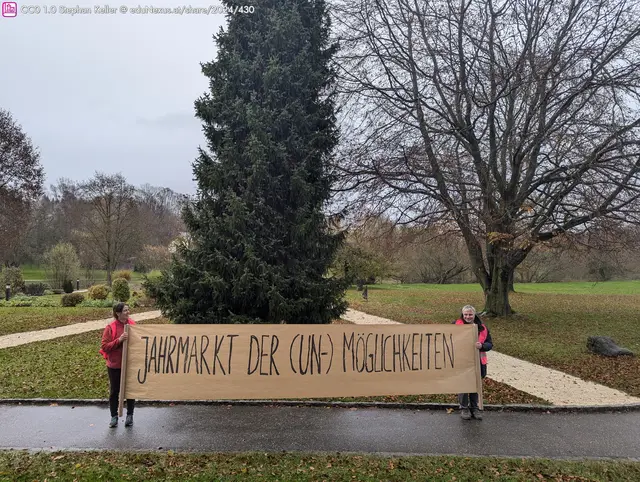 Zwei Personen halten ein großes Banner mit der Aufschrift „JAHRMARKT DER (UN-) MÖGLICHKEITEN“ vor einem großen Baum in einem Park.