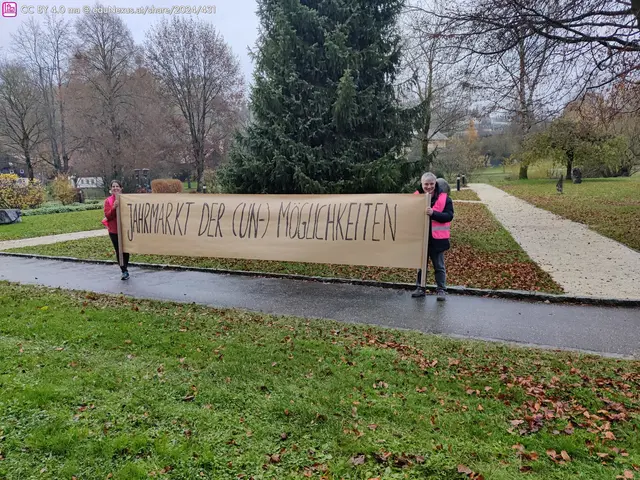 Zwei Personen halten ein großes Banner mit der Aufschrift „Jahrmarkt der (Un-)Möglichkeiten“ in einem Park. Im Hintergrund sind Bäume und ein schmaler Weg sichtbar.