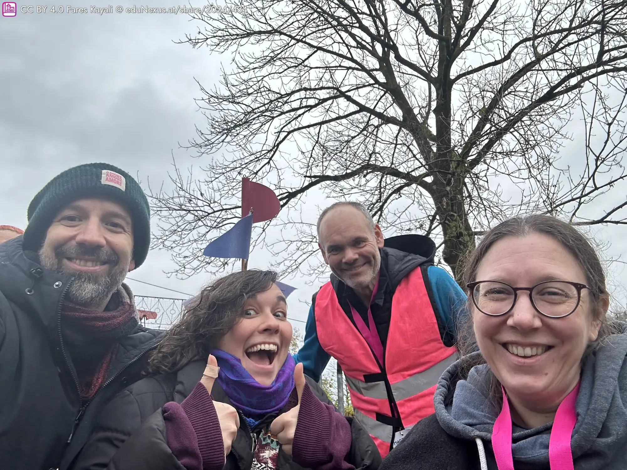 Vier Personen posieren in einem Selfie, lächeln und zeigen Daumen hoch. Im Hintergrund ein Baum mit grauem Himmel und bunte Fahnen.