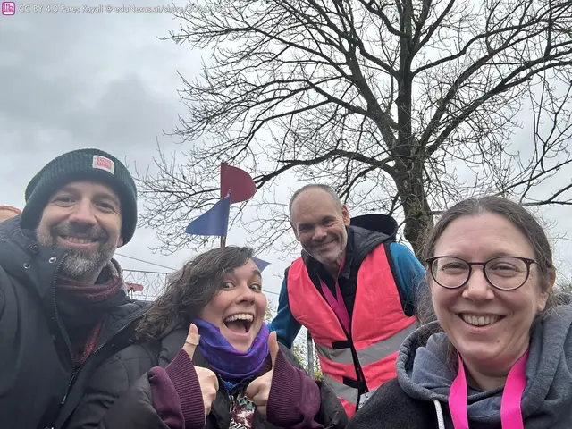 Vier Personen posieren in einem Selfie, lächeln und zeigen Daumen hoch. Im Hintergrund ein Baum mit grauem Himmel und bunte Fahnen.