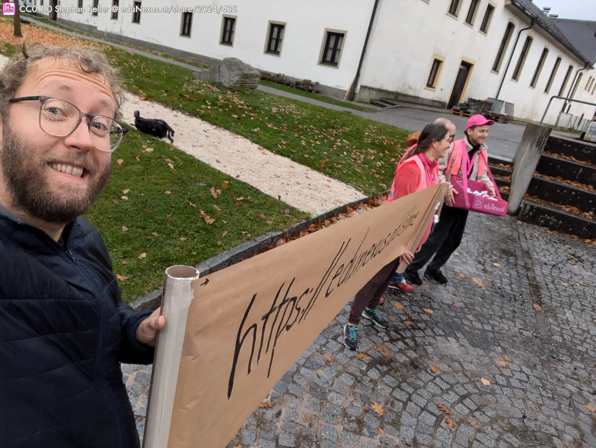 Mann mit Brille und lockigem Haar hält ein Schild, während drei Personen in pinken Shirts daneben stehen. Im Hintergrund ein Gebäude und ein Weg mit Gras.