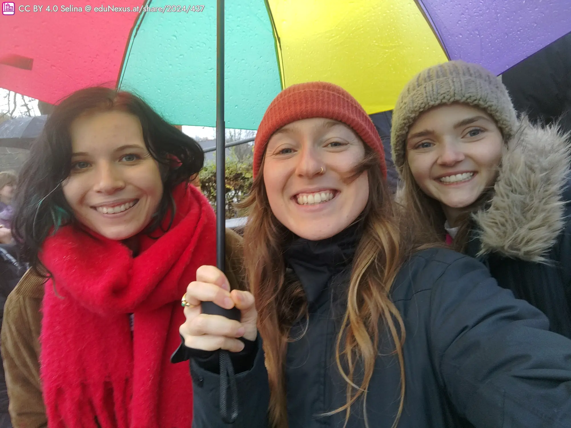 Drei Frauen lächeln unter einem bunten Regenschirm. Die linke trägt einen roten Schal, die mittlere eine rote Mütze und die rechte einen grauen Hut.