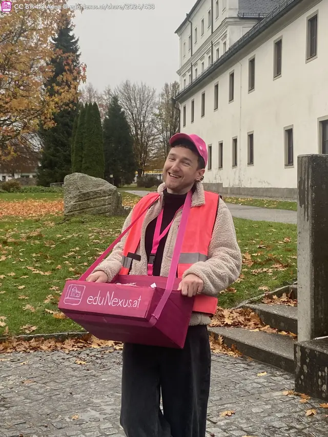 Junger Mann mit lächelndem Gesicht, trägt eine pinke Mütze und eine orange Weste. Er hält eine pinke Box mit der Aufschrift "eduNexus.at". Herbstliche Umgebung mit gefallenen Blättern.