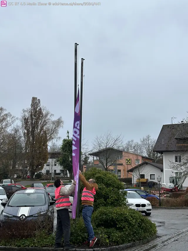 Zwei Personen in pinken Westen ziehen an einem großen, lila Banner, während es an einem Fahnenmast hängt. Im Hintergrund sind Autos und ein Wohngebäude zu sehen.