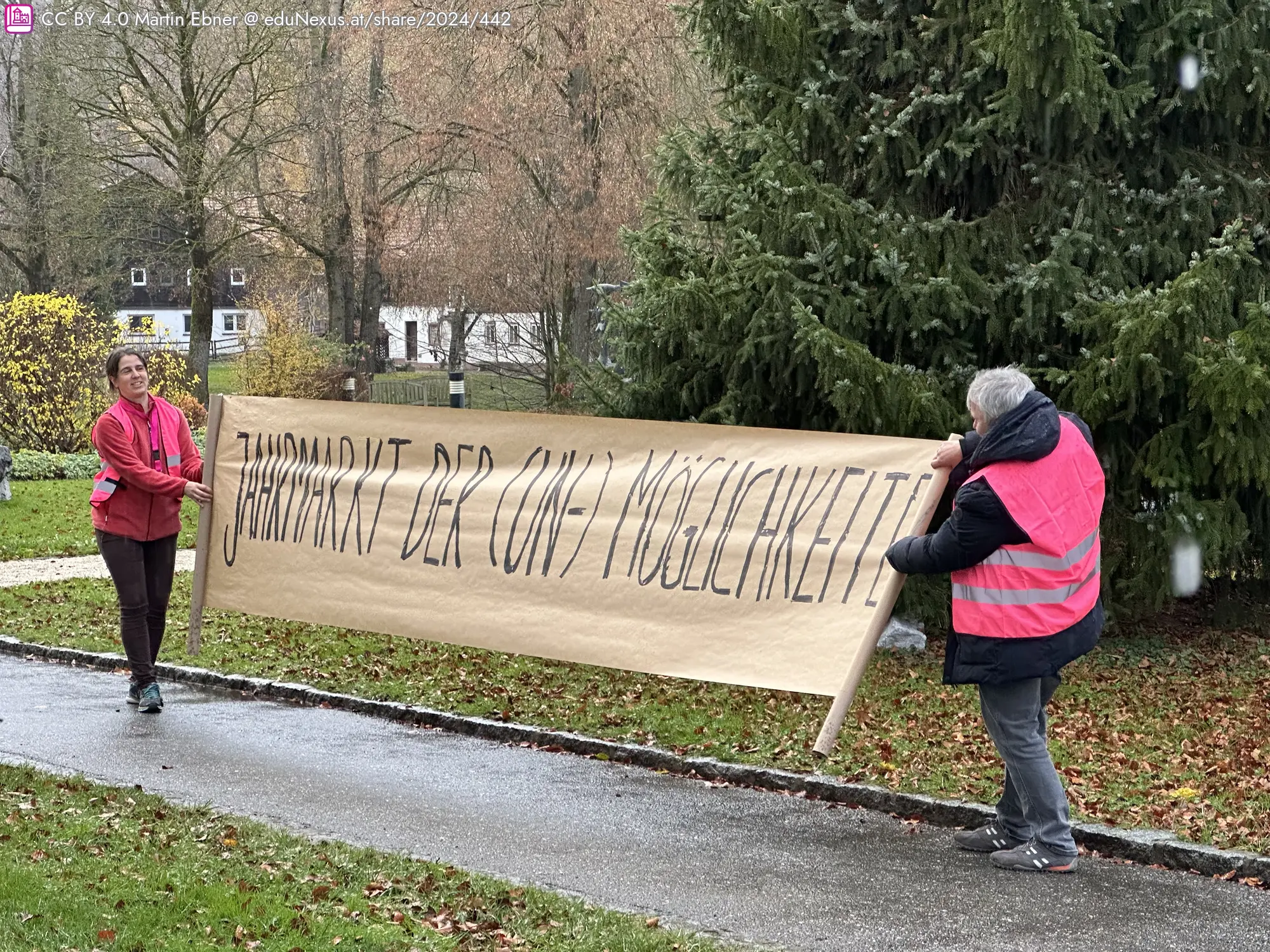 Zwei Personen tragen ein großes Banner mit der Aufschrift „Jahmark der Un- Möglichkeiten“ in einem Park.