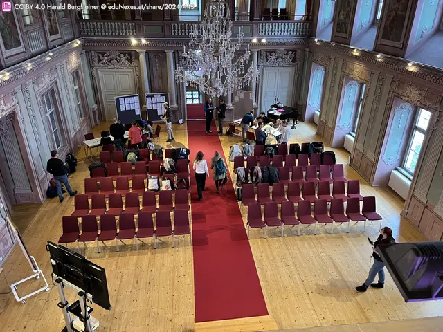 Eine große, elegante Halle mit einem roten Teppich in der Mitte, umgeben von Reihen roter Stühle. Menschen stehen und sitzen, während ein Kristalllüster von der Decke hängt.