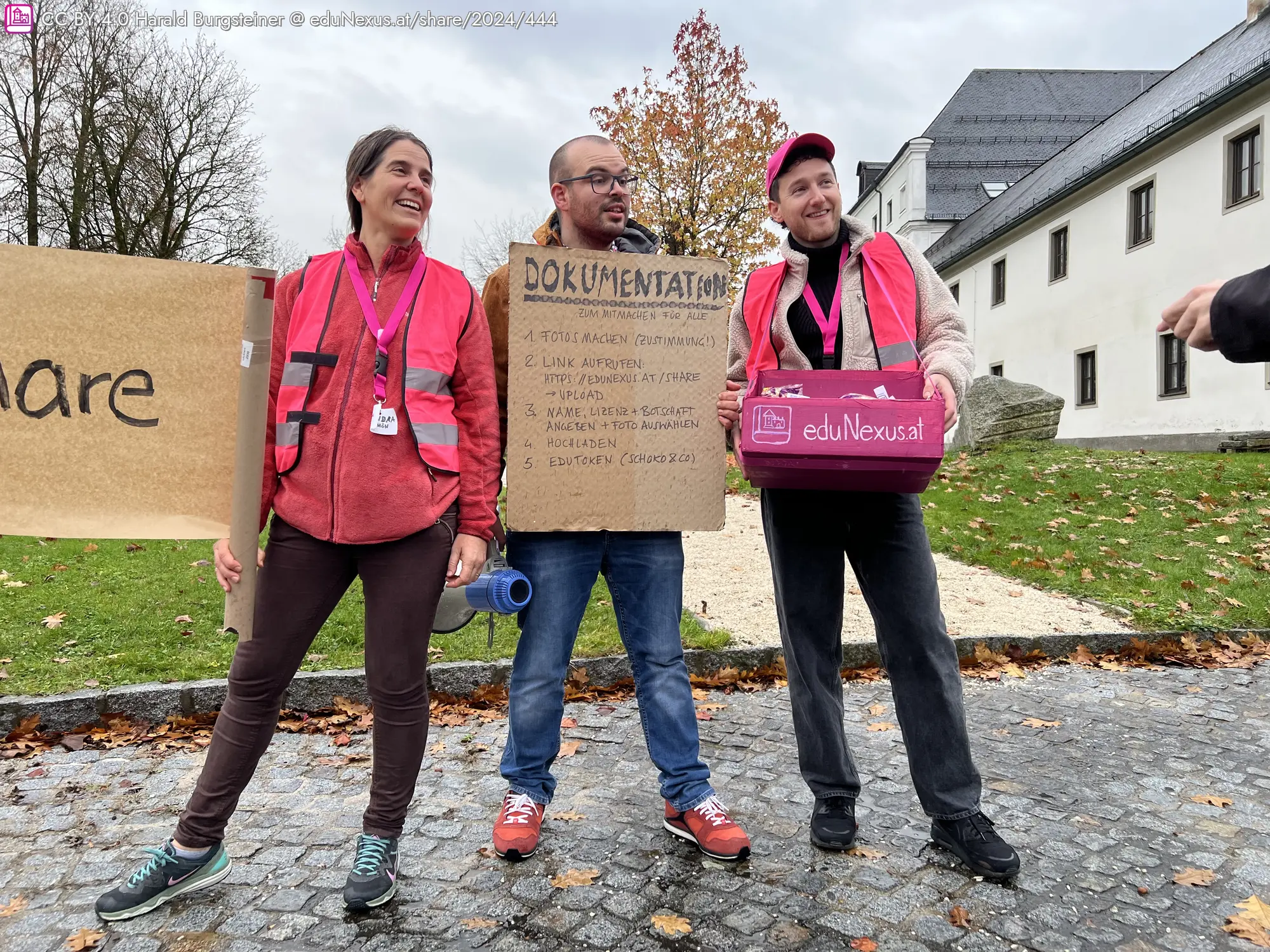 Drei Personen stehen im Freien, tragen pinkfarbene Westen und halten Schilder. Eine Person hält ein großes, handgeschriebenes Schild mit der Aufschrift „DOKUMENTATION“. Eine andere Person hält eine Kiste mit der Aufschrift „eduNexus.at“. Im Hintergrund sind Bäume und ein Gebäude sichtbar.