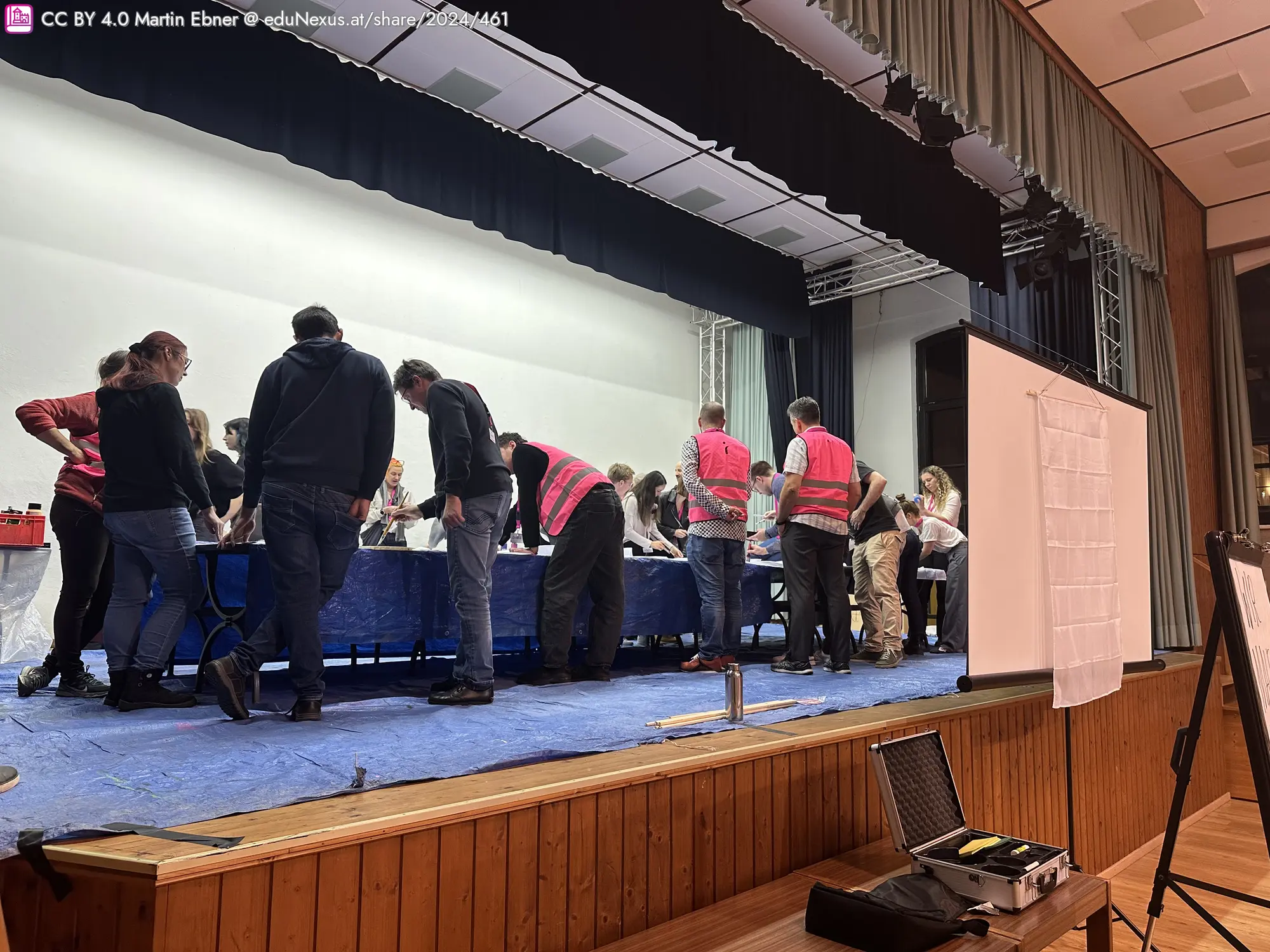 Menschen in rot-weißen Westen stehen um einen Tisch, auf dem Materialien liegen. Im Hintergrund eine Bühne mit Vorhängen, an der Seite ein Flipchart und ein Koffer.