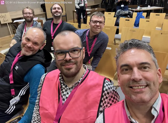 Gruppe von sechs Männern in pinken Westen, lächelnd in einem Raum mit Holzdielen und Stühlen im Hintergrund.