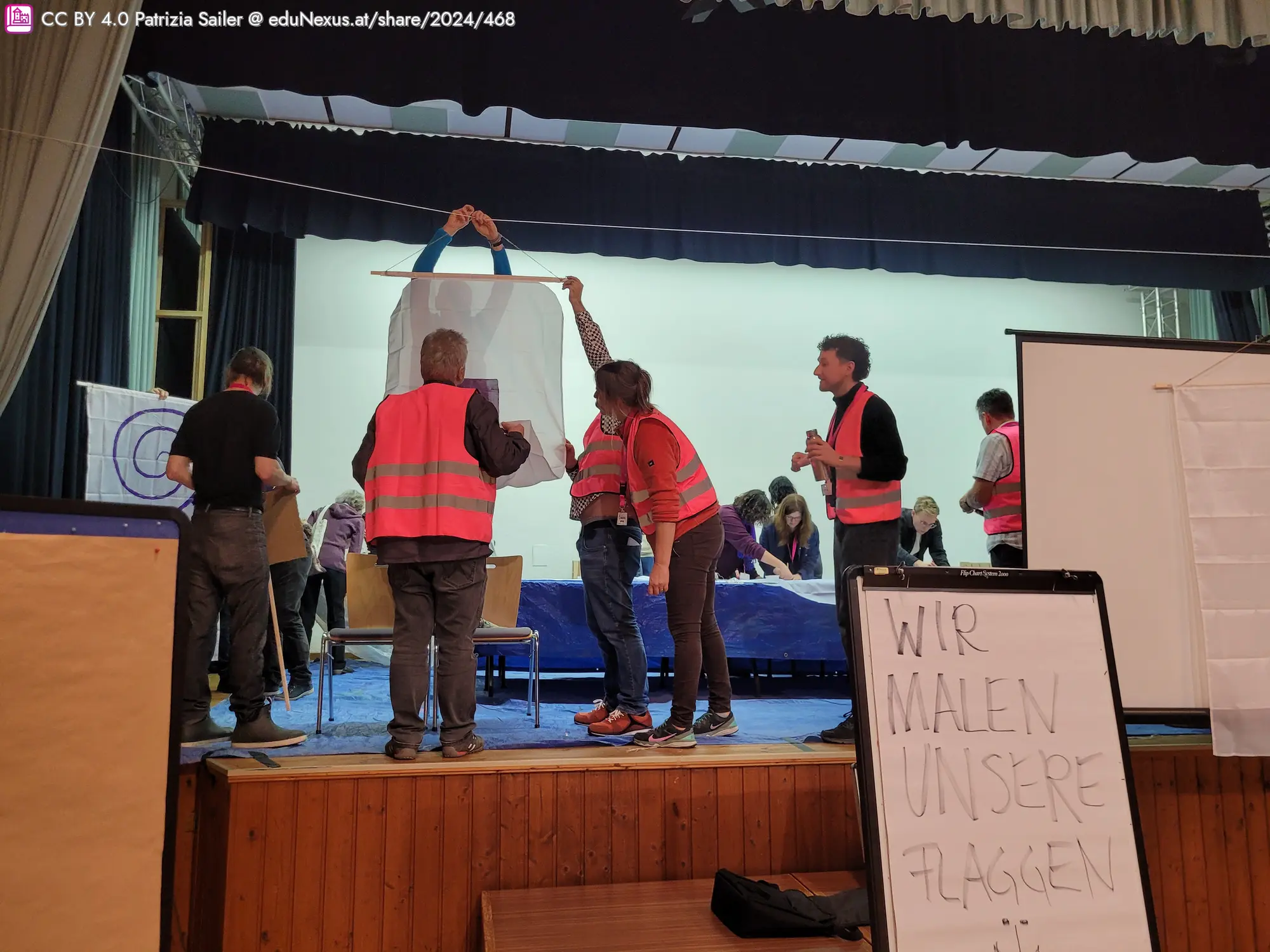 Menschen in Warnwesten arbeiten gemeinsam auf einer Bühne, um ein großes weißes Tuch zu halten. Im Vordergrund steht ein Schild mit der Aufschrift „WIR MALEN UNSERE FLAGGEN“.