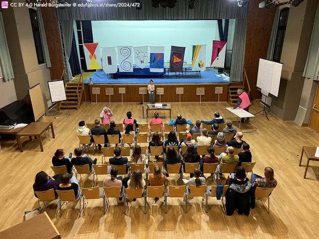 Eine Bühne mit bunten Bannern im Hintergrund, eine Person steht vorne und spricht zu einem Publikum, das auf Stühlen sitzt. Tische und Flipcharts sind im Raum verteilt.