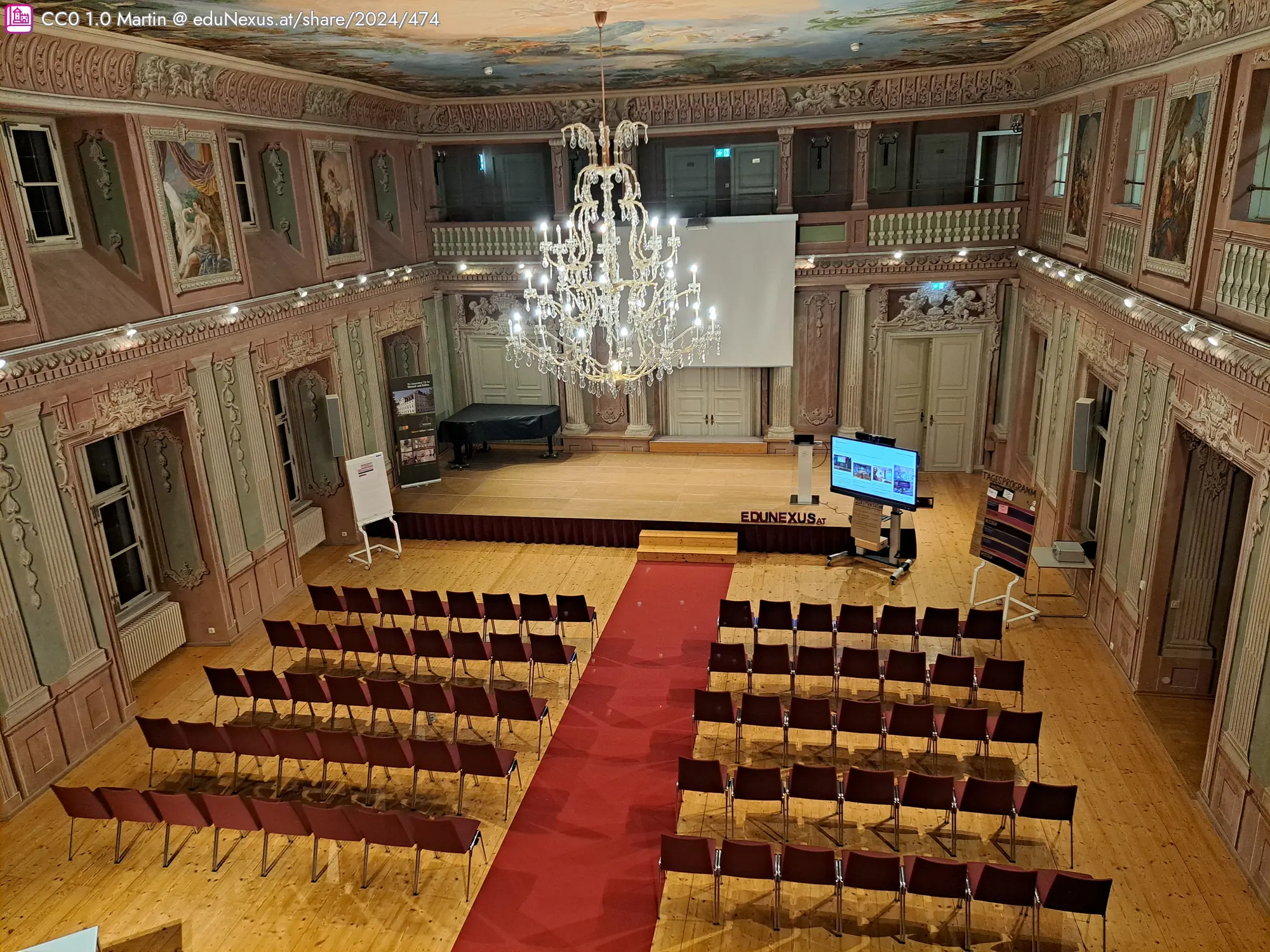 Ein eleganter Saal mit einem Kristalllüster, einer roten Teppichmitte und Reihen von Stühlen. Eine Leinwand und ein Monitor sind auf der Bühne platziert. Dekorative Wandmalereien und große Fenster sind sichtbar.