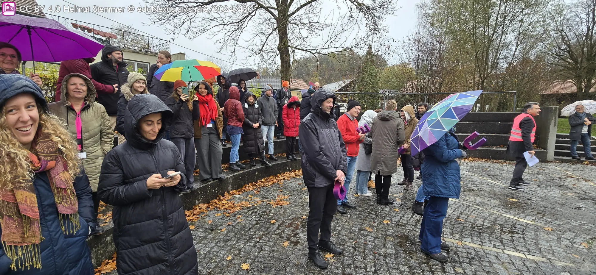 Die Teilnehmer stehen im (zarten) Regen, tragen tw. Regenmäntel und halten Regenschirme in verschiedenen Farben. Einige schauen auf ihr Handy, andere beobachten was sich so abseits des Bildinhaltes abspielt. Herbstliche Bäume im Hintergrund.