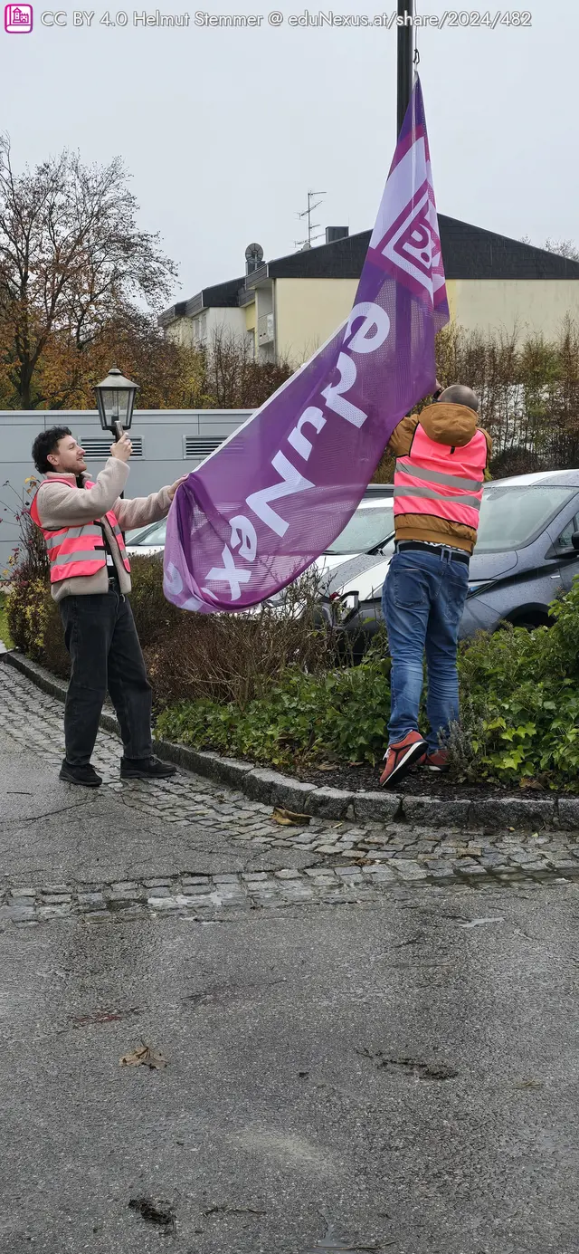 Zwei Personen in pinken Westen halten ein großes, lila Banner mit der Aufschrift eduNEXUS. Eine Person steht auf einer gepflasterten Fläche, während die andere an einem Pfosten zieht. Im Hintergrund sind einige (unscharf) Autos und Bäume im spätherbstlichen Outfit sichtbar.