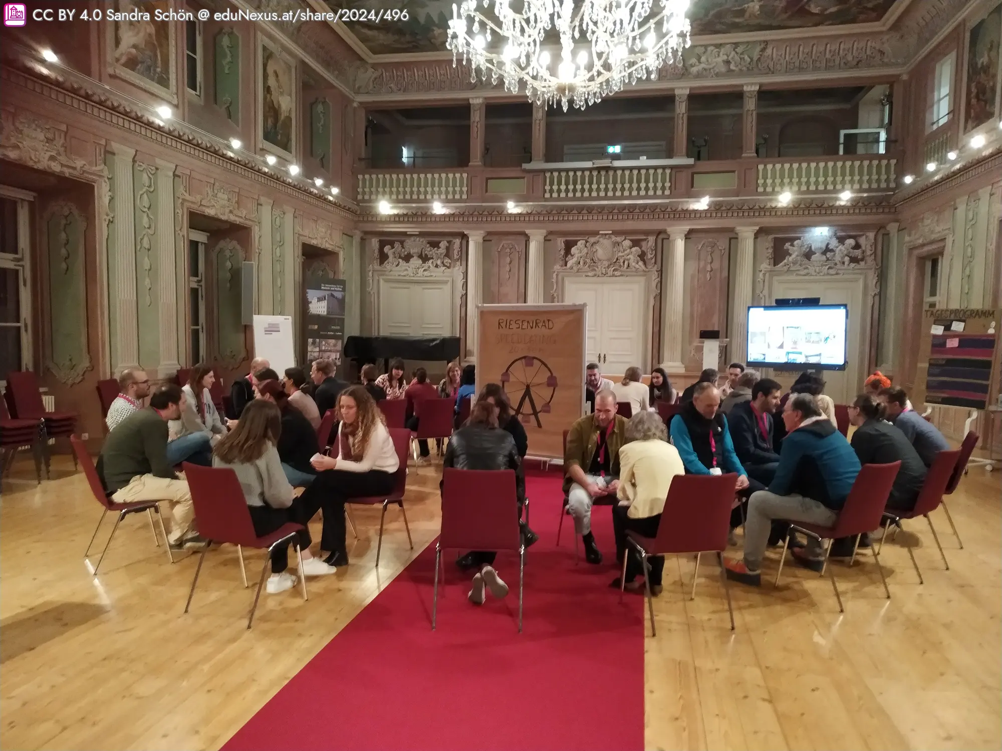 Großer Saal mit eleganter Dekoration, zwei Gruppen von Personen, die miteinander sprechen. In der Mitte steht eine Holztafel mit dem Wort „Riesenrad Speeddating“.