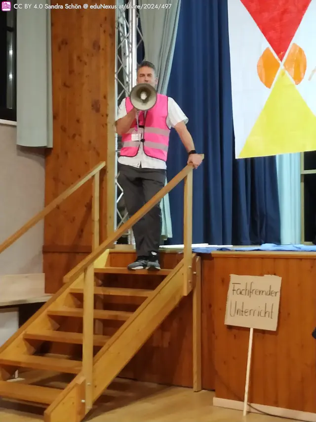 Mann mit Megafon in pinker Weste steht auf einer Holztreppe. Im Hintergrund ein Banner mit roten und gelben Symbolen sowie ein Schild mit der Aufschrift „Fachfremder Unterricht“.