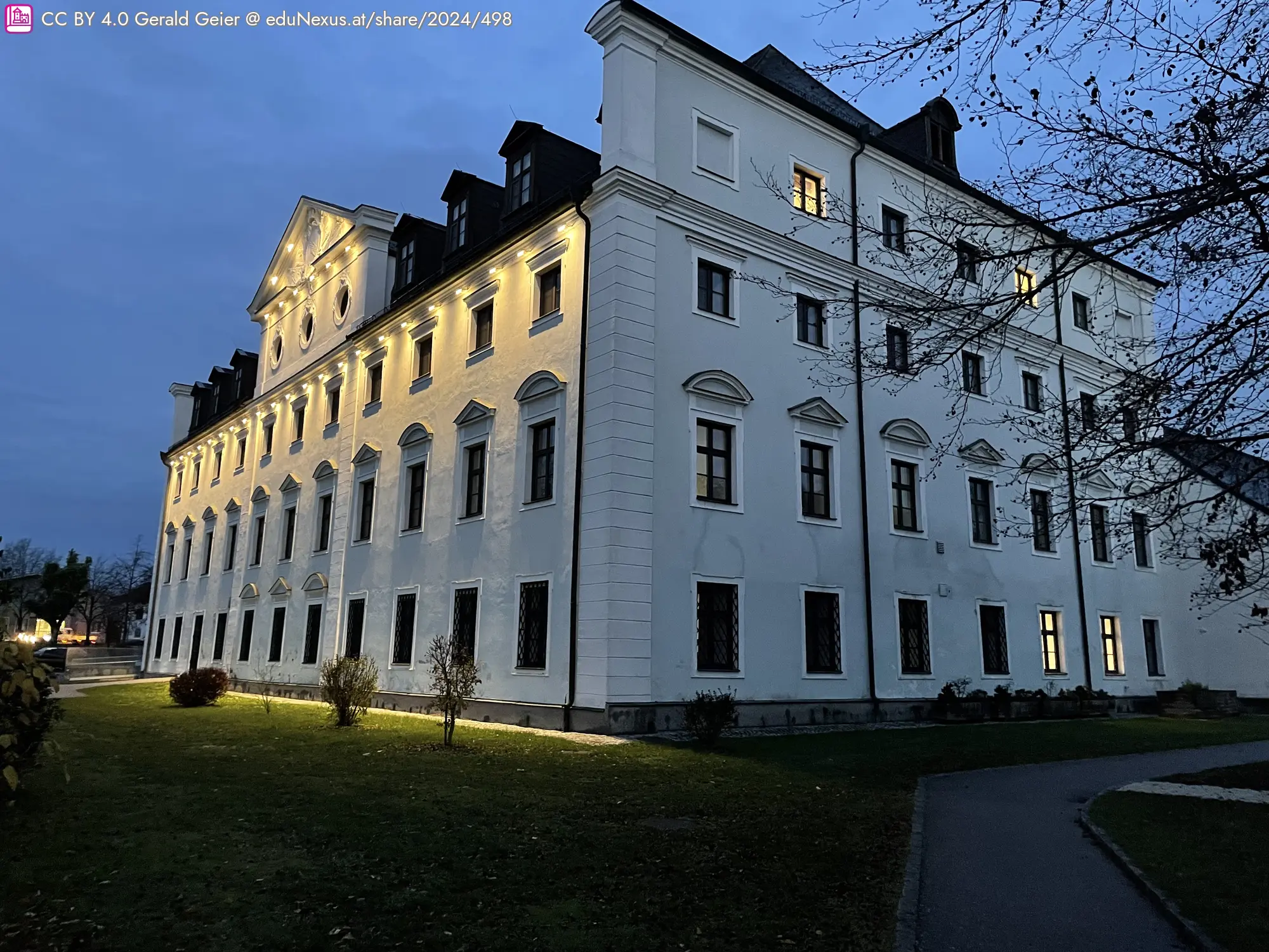 Schloss mit mehreren Fenstern, beleuchtet, umgeben von Bäumen und einem gepflegten Garten, im Morgenlicht.