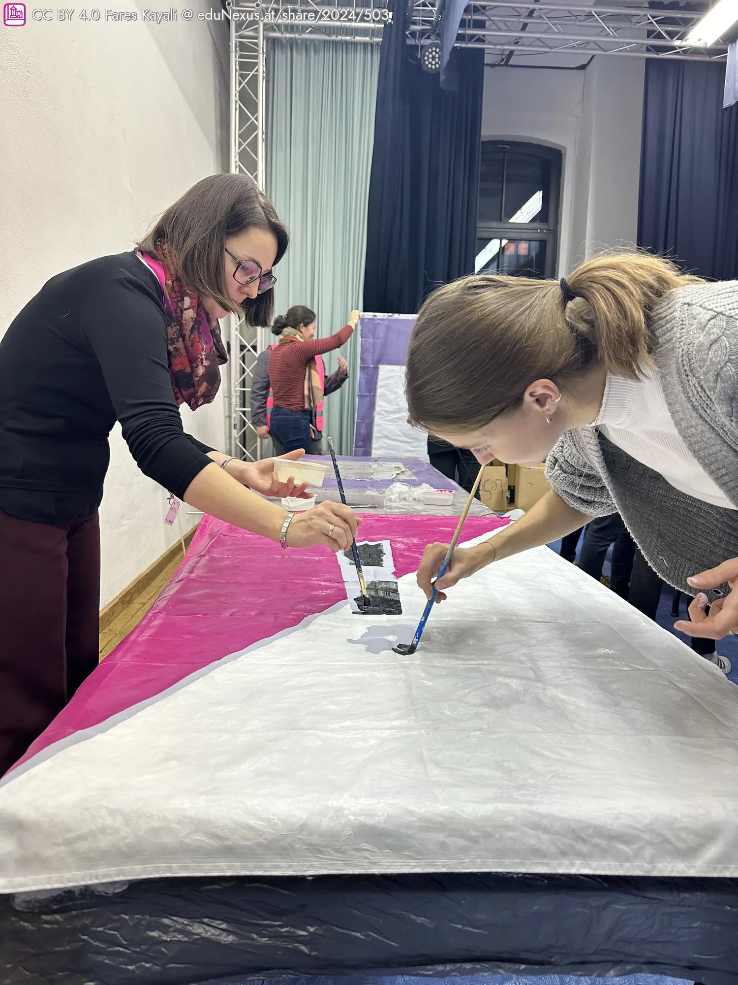 Zwei Frauen malen mit Pinseln auf einem großen, bunten Stoff. Eine Frau in Schwarz und eine in Grau konzentrieren sich auf ihre Arbeit. Im Hintergrund sind weitere Personen sichtbar.