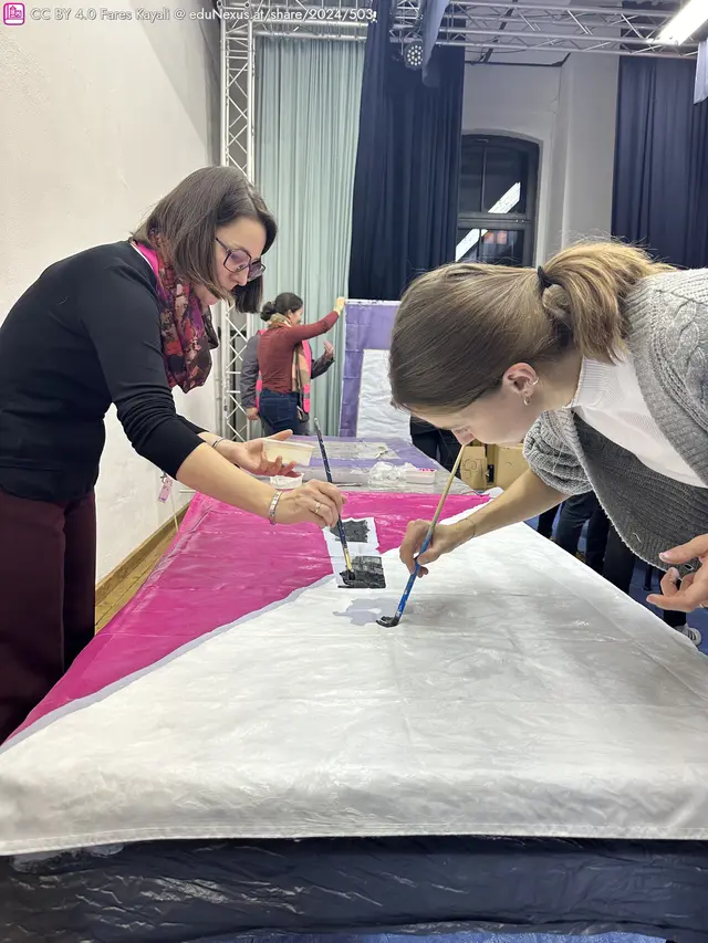 Zwei Frauen malen mit Pinseln auf einem großen, bunten Stoff. Eine Frau in Schwarz und eine in Grau konzentrieren sich auf ihre Arbeit. Im Hintergrund sind weitere Personen sichtbar.