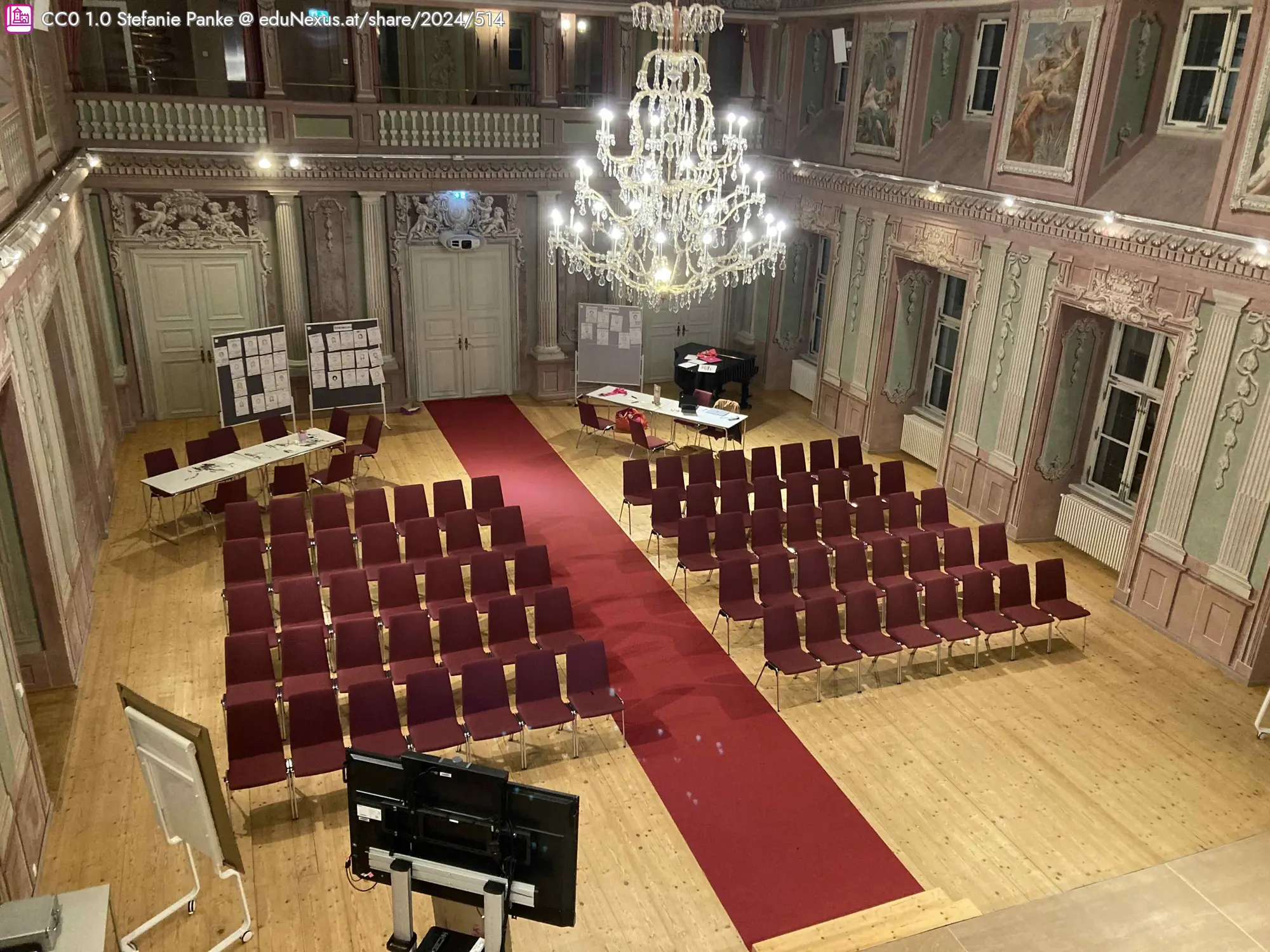 Veranstaltungsraum mit rot ausgestatteten Stühlen, rotem Teppich, einem Kronleuchter und einem Tisch mit Materialien im Hintergrund.