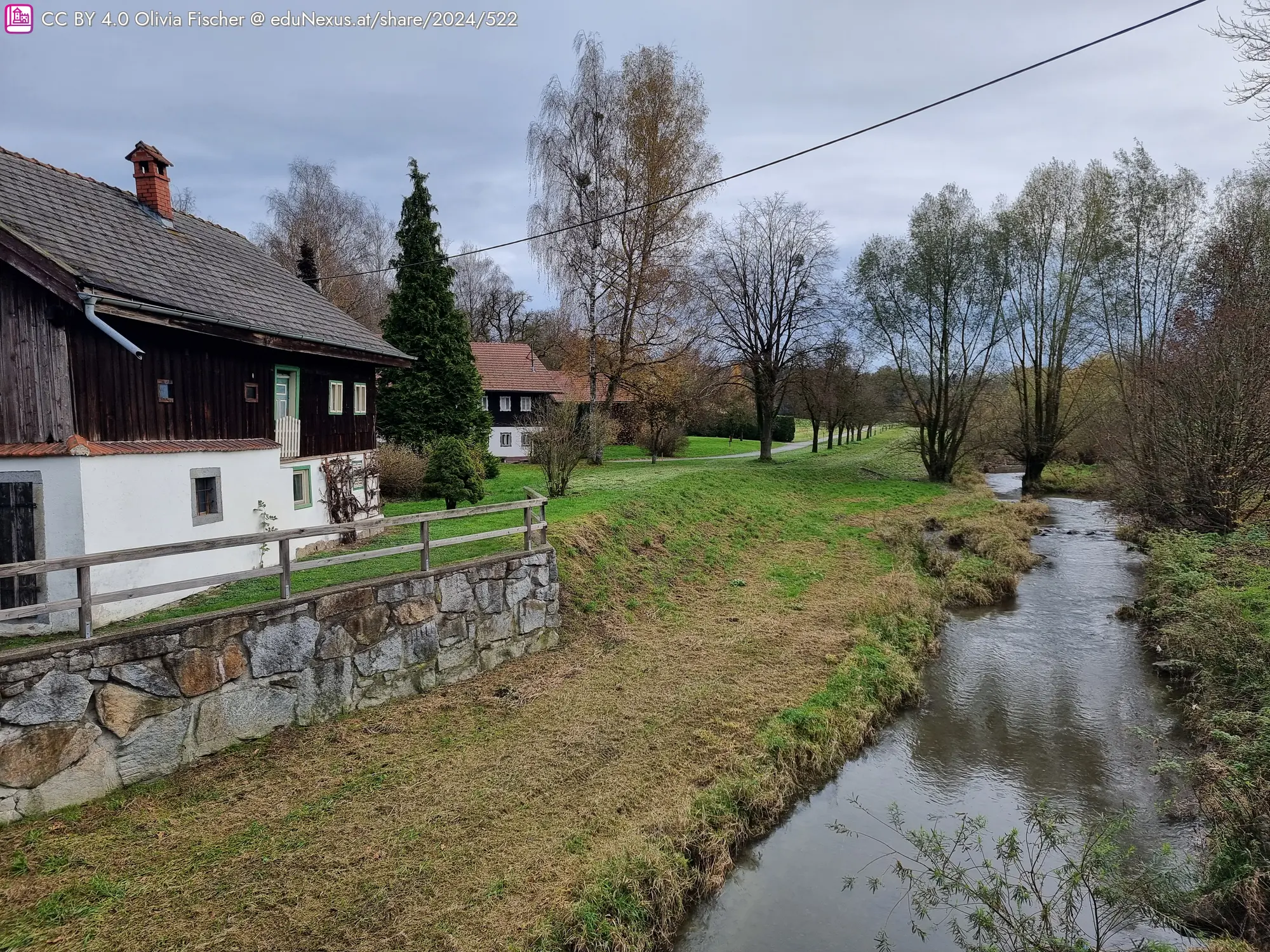 Zwei Häuser am Ufer eines kleinen Baches, umgeben von Bäumen und Wiese, unter einem bewölkten Himmel.