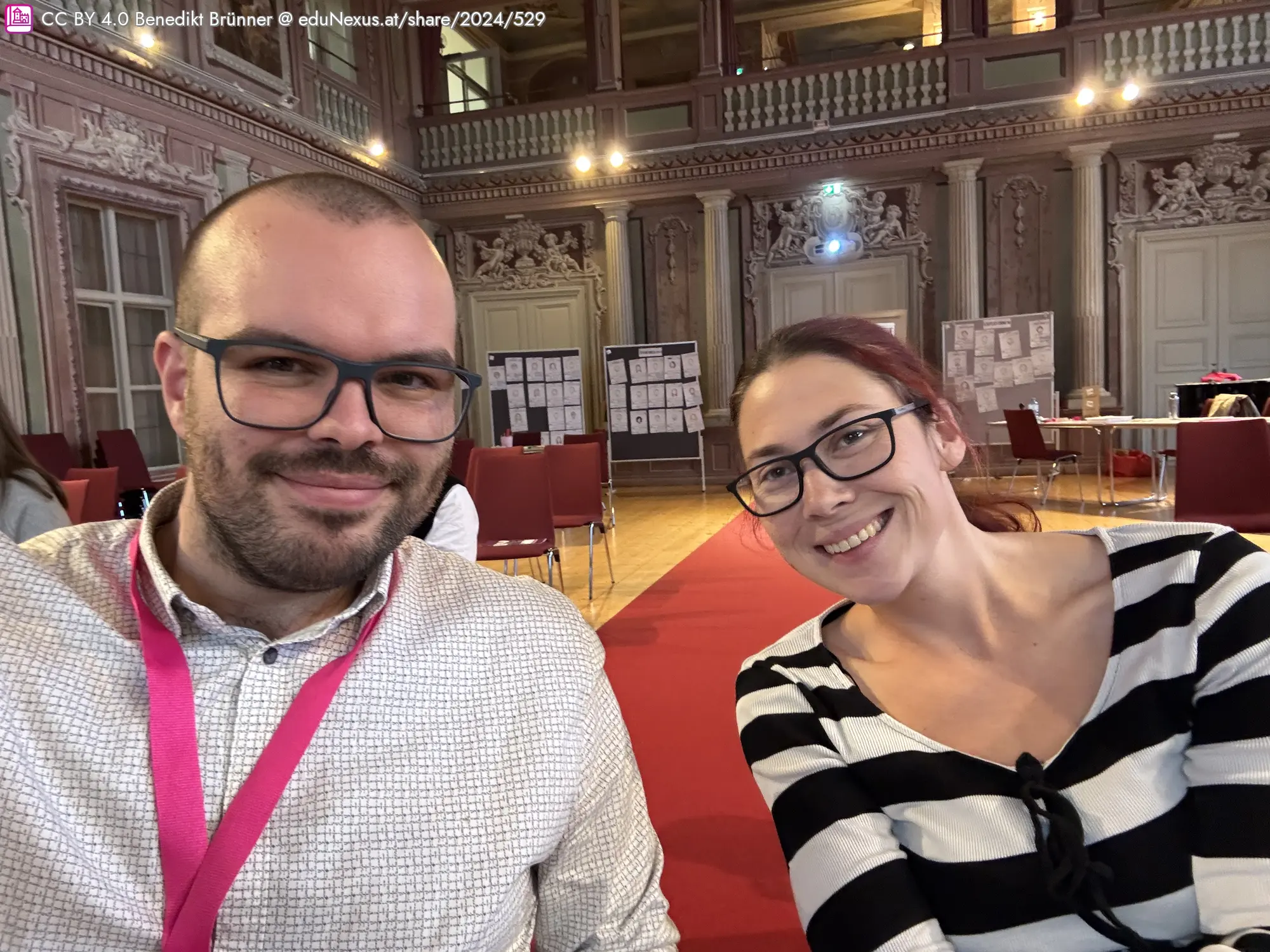 Ein Mann und eine Frau lächeln in einem großen, lichtdurchfluteten Raum mit roten Teppich und Stühlen im Hintergrund.