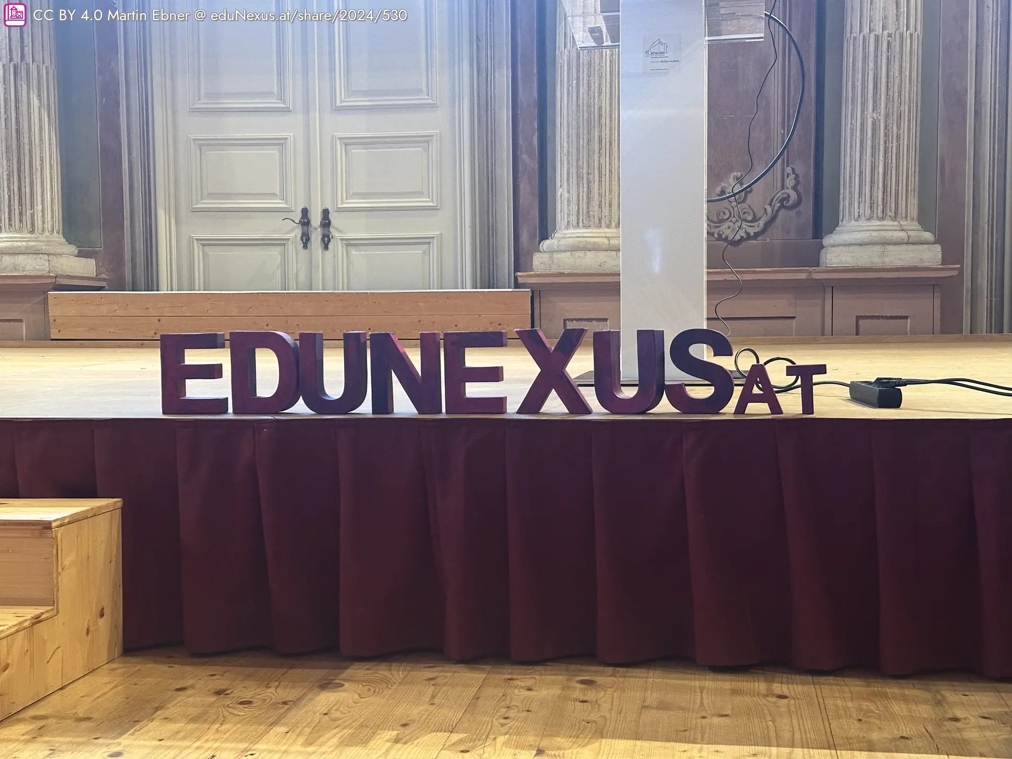 Das Wort „EDUNEXUS“ in großen, bunten Buchstaben auf einer Bühne mit rotem Vorhang und einer weißen Säule im Hintergrund.