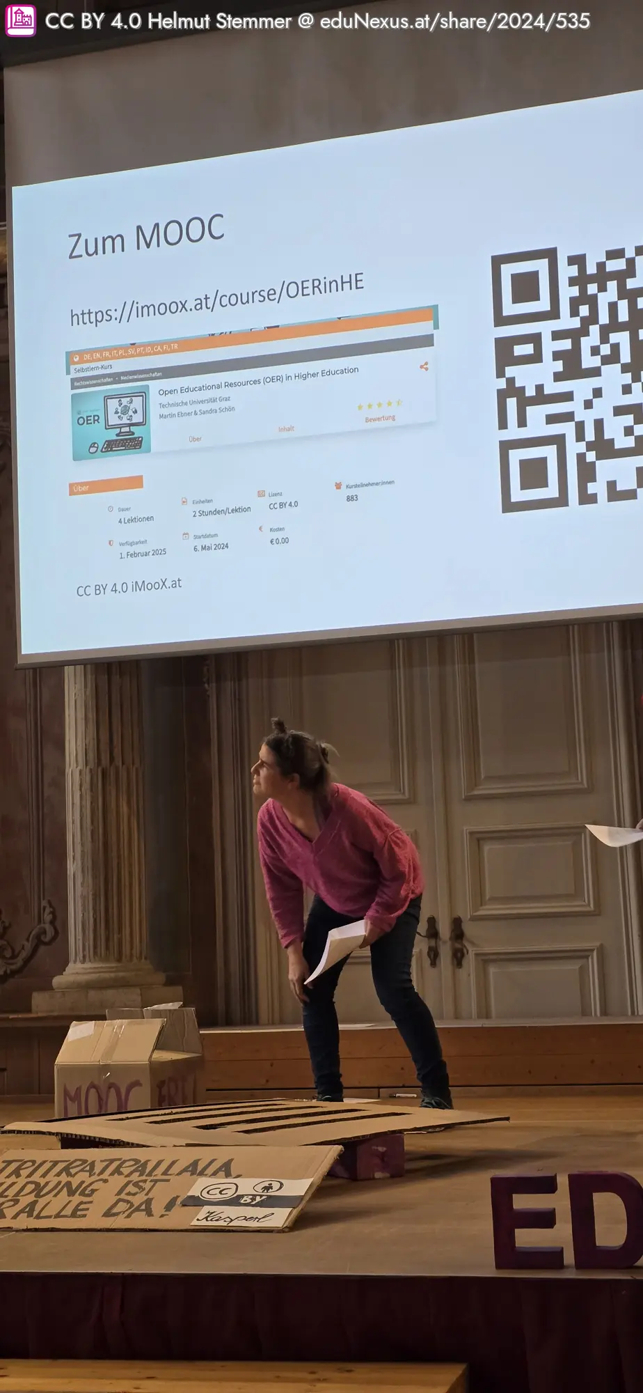 Eine Frau in einem rosa Oberteil steht auf einer Bühne mit einer Präsentation im Hintergrund, die einen QR-Code und Informationen zu einem MOOC zeigt. Auf dem Boden liegen beschriftete Pappschilder und Kartons.