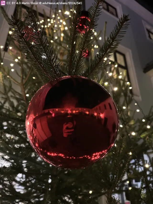 Rote Weihnachtsbaumkugel hängt an einem Tannenzweig, mit Lichtern im Hintergrund.