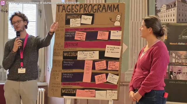 Ein Mann mit Brille und Locken präsentiert ein Tagesprogramm auf einem großen Plakat. Neben ihm steht eine Frau in einem roten Pullover. Das Plakat enthält verschiedene Zeitblöcke und Aktivitäten, die mit bunten Zetteln beschrieben sind.