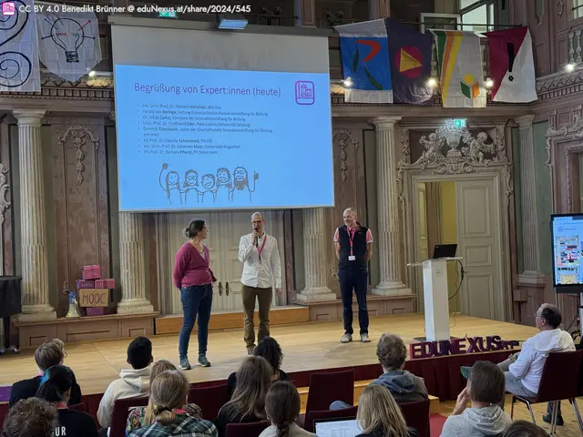 Drei Personen stehen auf einer Bühne vor einer Präsentation. Im Hintergrund ist ein großes Display mit Text und Illustrationen. Im Publikum sind Teilnehmer zu sehen. Bunte Flaggen hängen an der Decke.