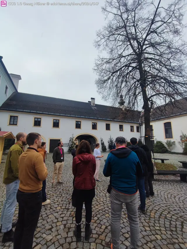 Gruppe von Menschen steht auf einem gepflasterten Platz vor einem weißen Gebäude mit Fensterläden. Ein Baum und einige Pflanzen sind im Hintergrund sichtbar.