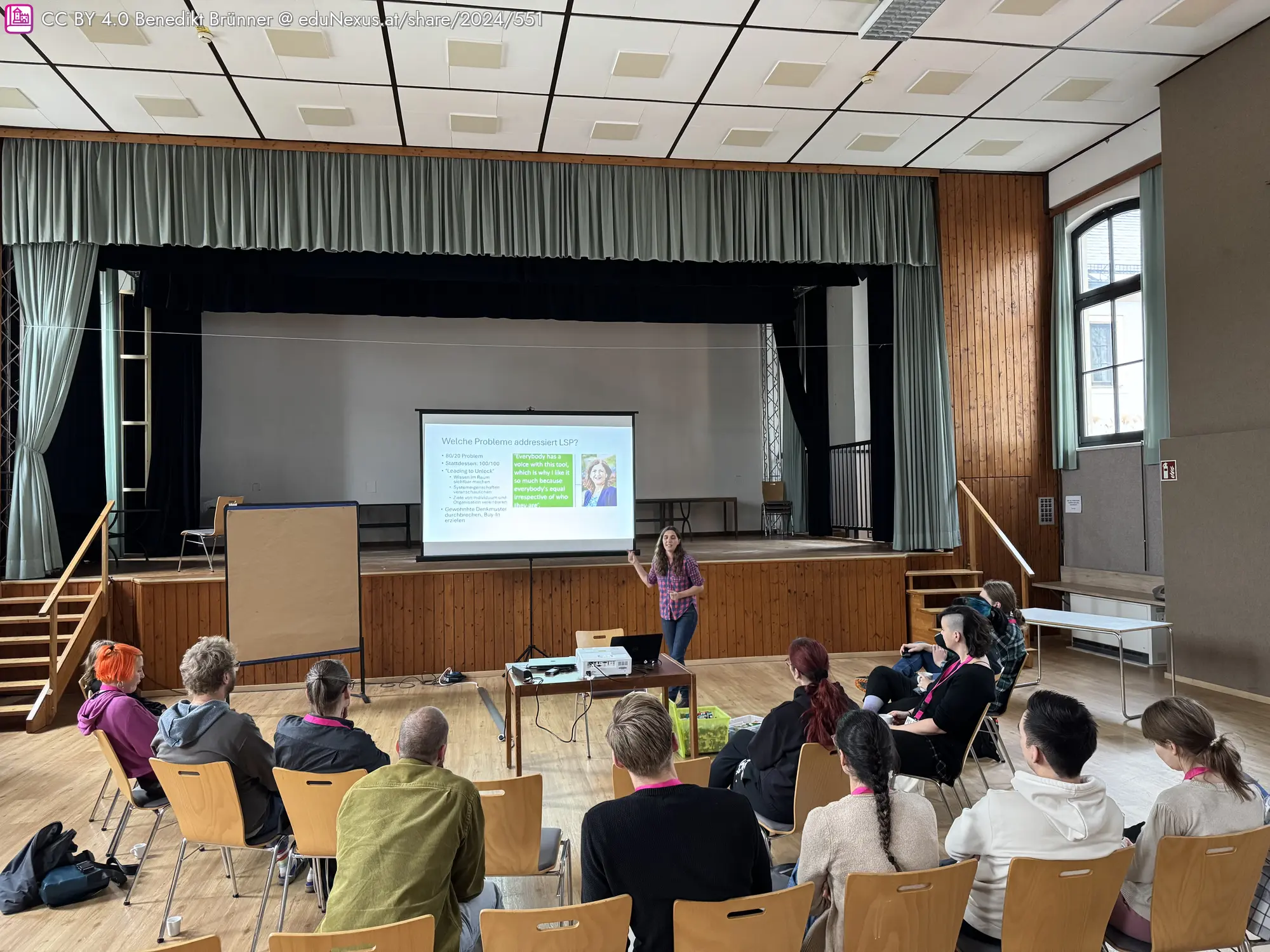 Eine Frau präsentiert vor einer Gruppe von Menschen in einem Saal. Auf einer Leinwand sind Informationen und ein Bild zu sehen. Stühle sind im Halbkreis angeordnet, und ein Flipchart steht neben der Präsentation.