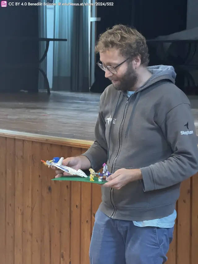 Mann mit Brille in grauem Hoodie hält eine grüne Platte mit LEGO-Figuren und einem Modell eines Flugzeugs. Holzverkleidung im Hintergrund.