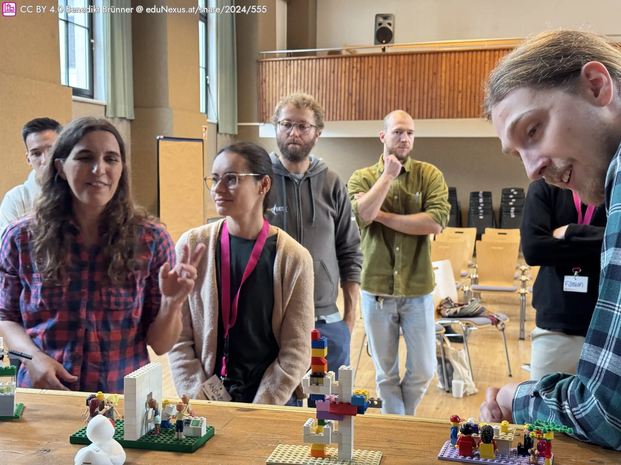 Eine Gruppe von fünf Personen steht am Rande der Bühne mit Lego-Modellen. Zwei Frauen diskutieren, während drei Männer aufmerksam zuhören.