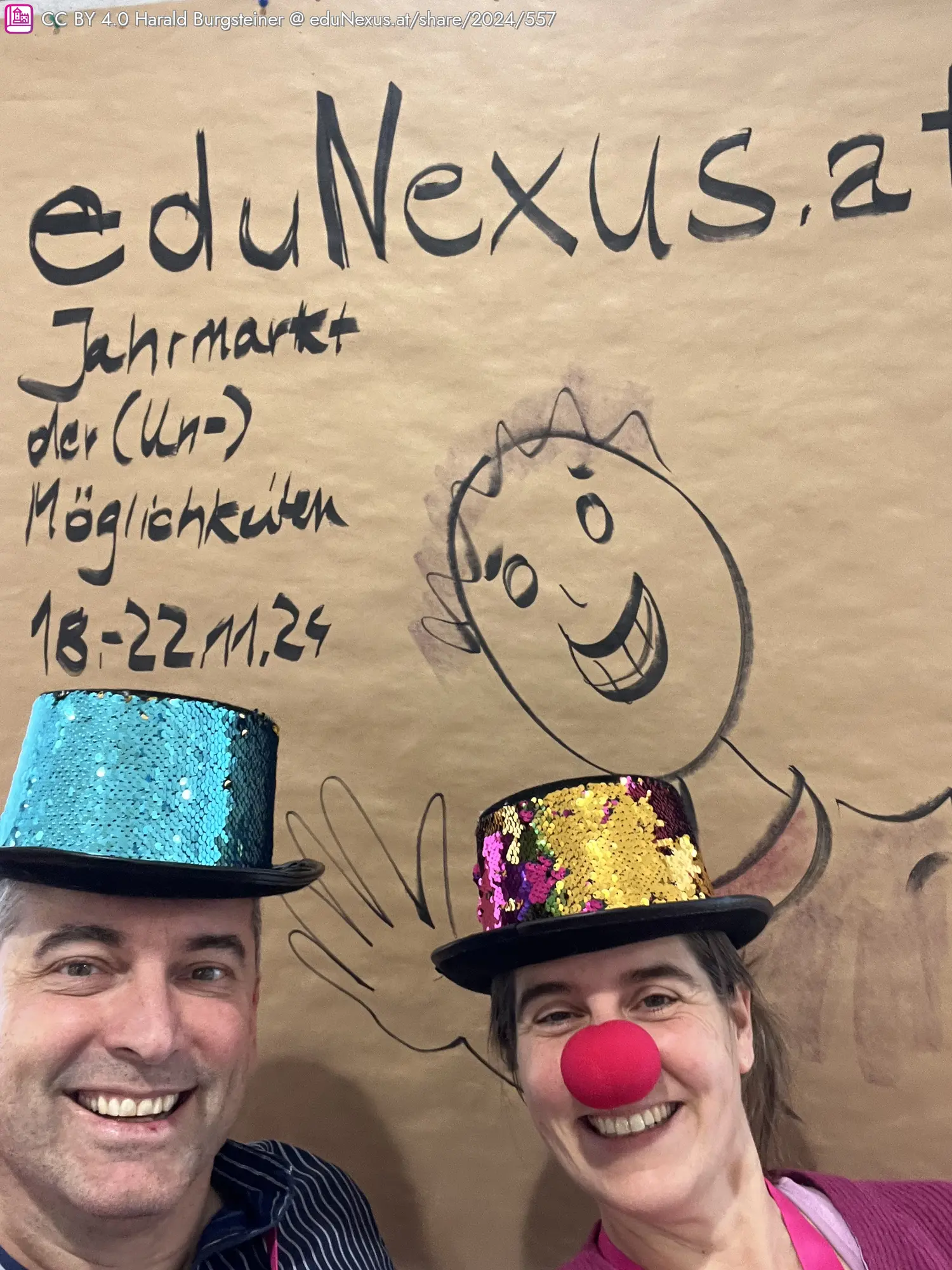 Selbstporträt von zwei Personen mit glitzernden Hüten, eine mit einer roten Nase, vor einem großen, braunen Papier mit dem Text „eduNexus.at Jahrmarkt der (Un-) Möglichkeiten 18-22.11.24“ und einer gezeichneten Figur.