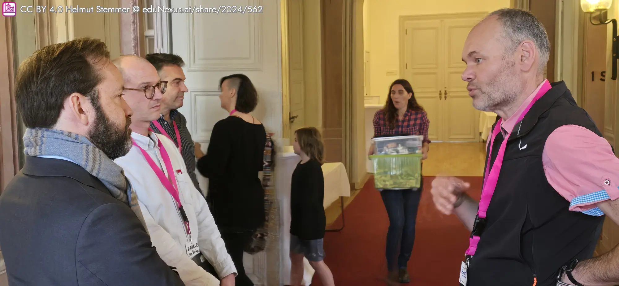 Fünf Personen im Gespräch, eine Frau im Hintergrund trägt eine Kiste. Alle tragen Ausweise mit pinken Lanyards.