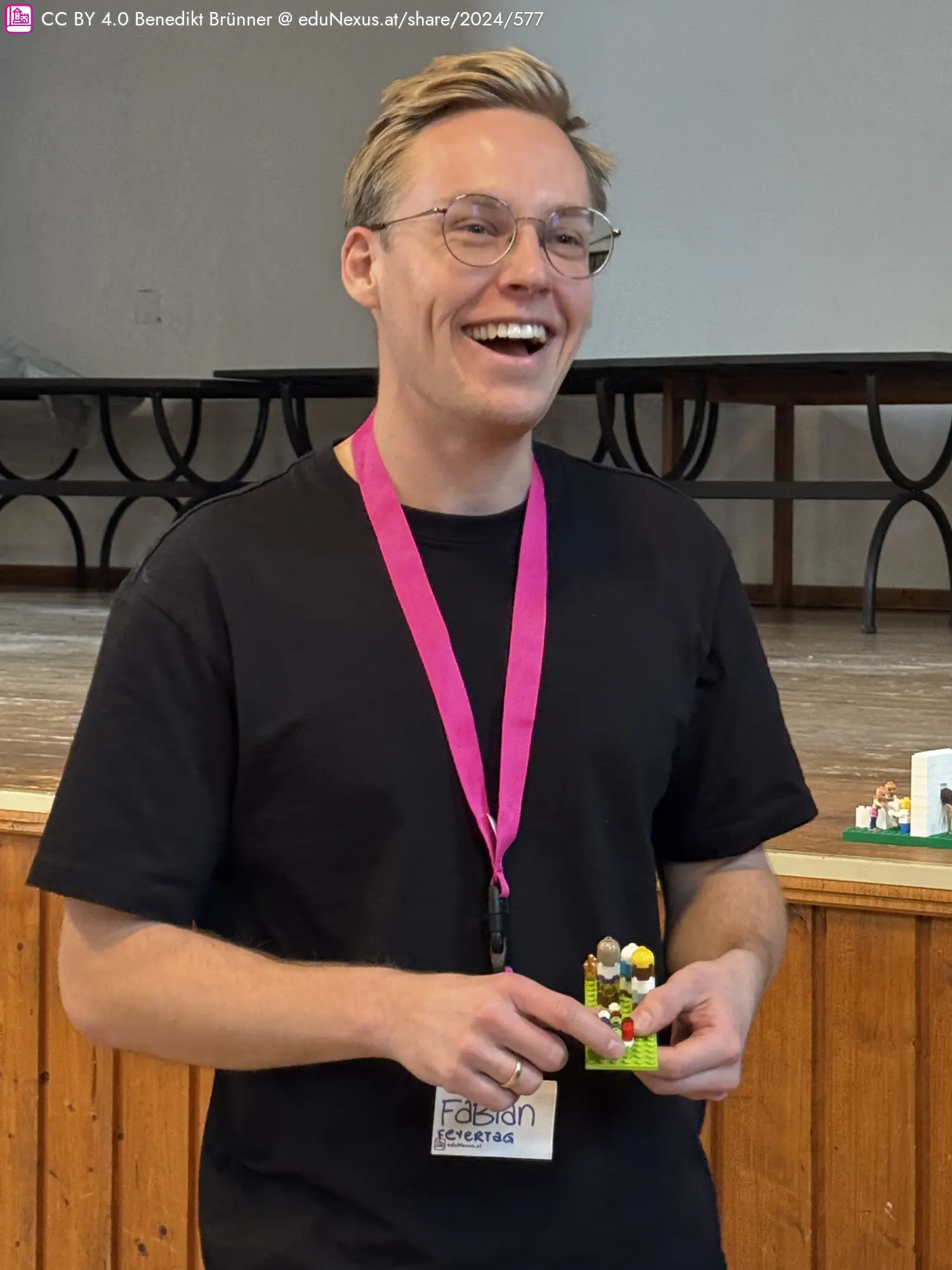 Lächelnder Mann mit Brille trägt ein schwarzes T-Shirt und eine pinke Lanyard, hält ein kleines Lego-Modell in der Hand.