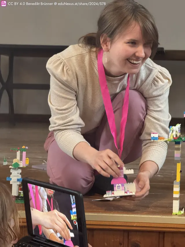 Eine lächelnde Frau mit einem rosa Band um den Hals sitzt auf dem Boden und baut mit bunten Bausteinen. Ein Tablet zeigt ihre Hände beim Arbeiten.