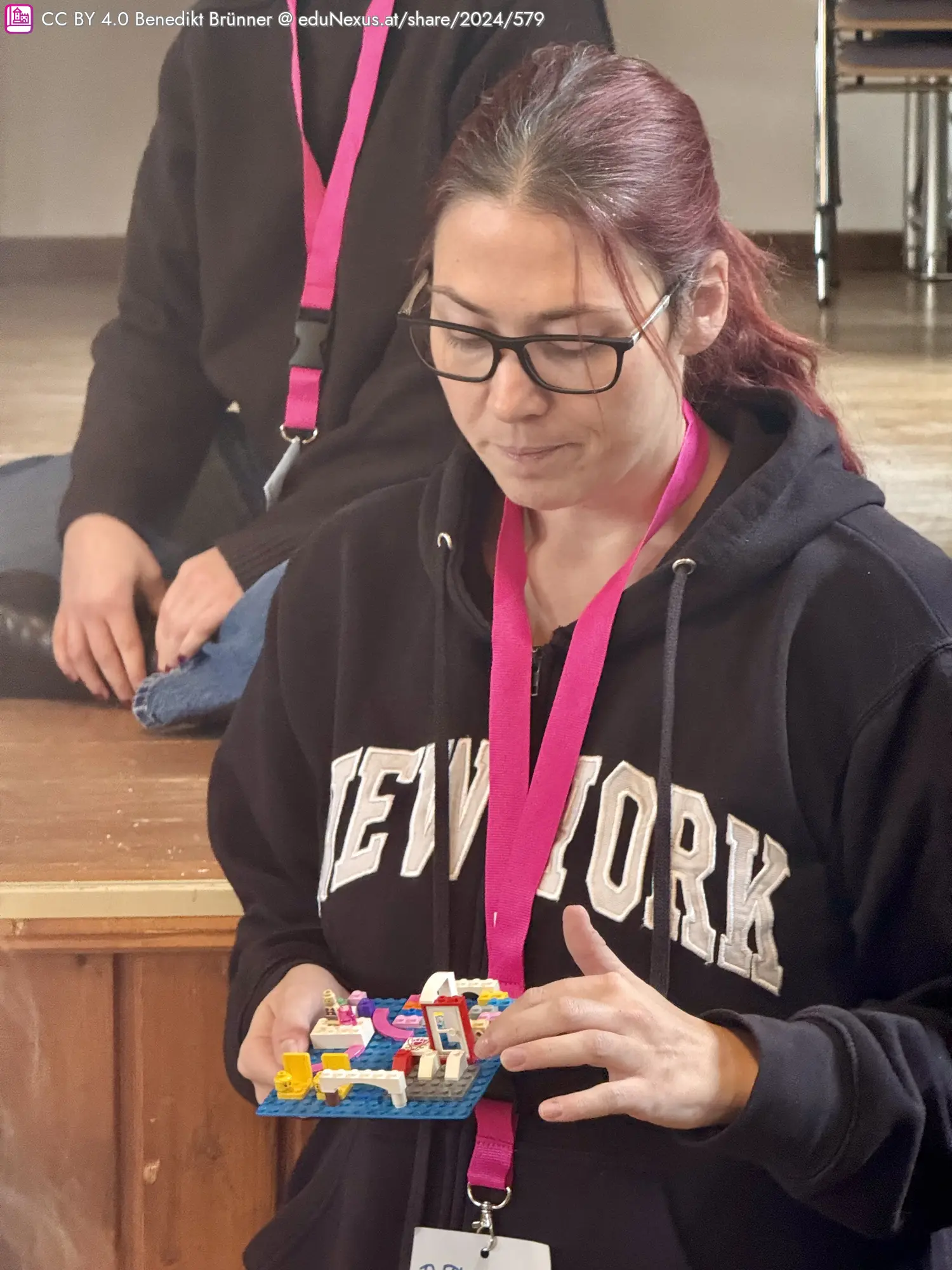 Eine Frau mit Brille und einem schwarzen Hoodie hält eine kleine, bunte Lego-Platte in der Hand. Sie trägt eine pinke Lanyard. Im Hintergrund sitzt eine weitere Person.