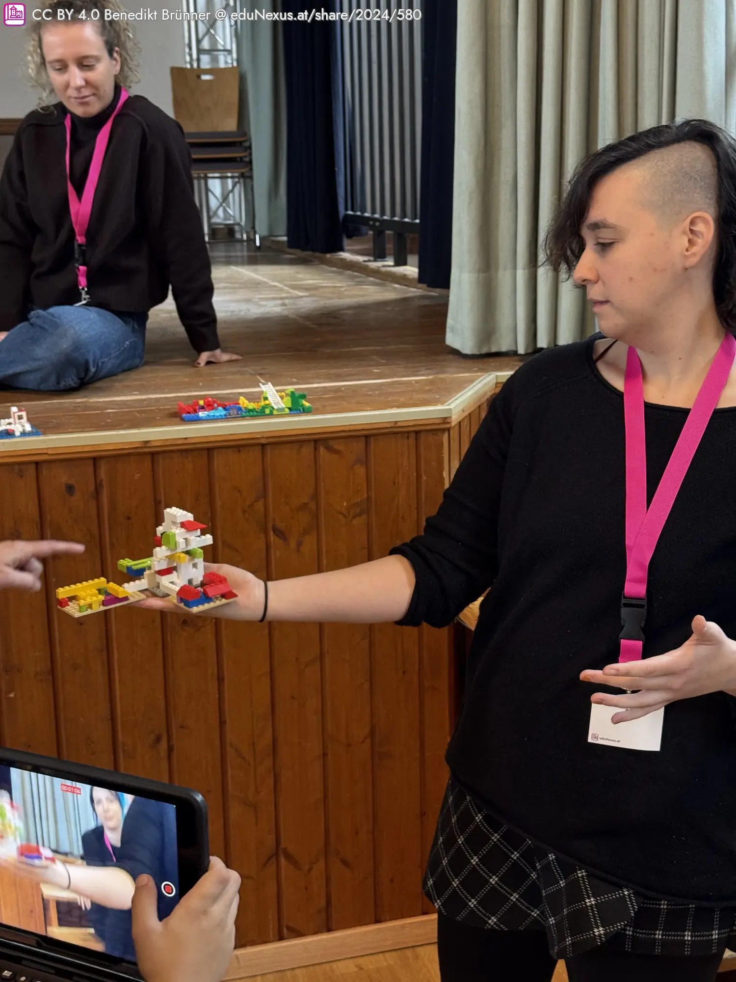 Eine Person mit kurzen Haaren hält ein Lego-Modell in der Hand. Im Hintergrund sitzt eine weitere Person.