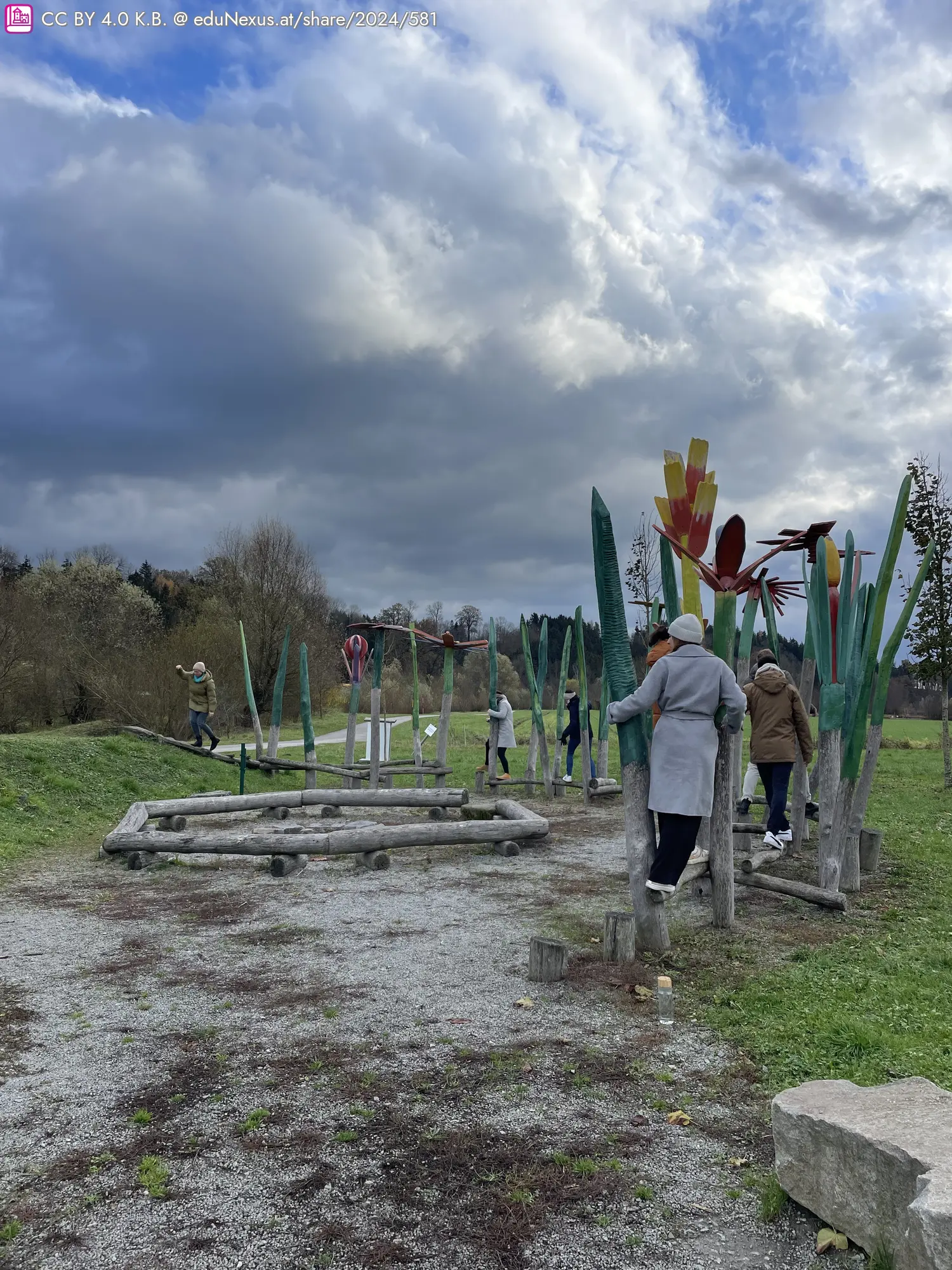 Spielplatz mit bunten, pflanzenartigen Skulpturen und Kindern, die spielen. Wolkiger Himmel im Hintergrund.