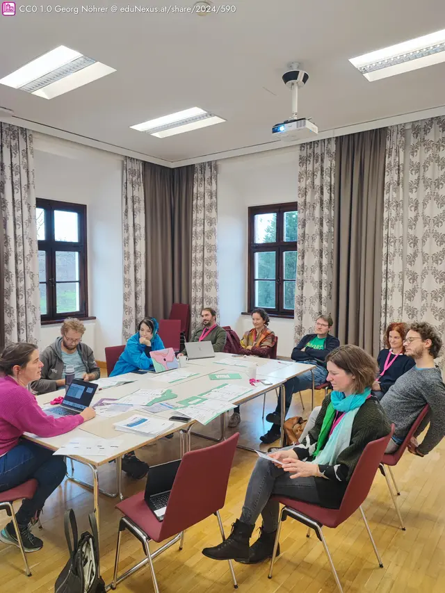 Ein Tisch in einem Raum mit großen Fenstern, umgeben von Stühlen. Eine Gruppe von Menschen hört einer Präsentation zu, einige nutzen Laptops und Tablets. Draperien mit Blumenmuster hängen an den Fenstern.