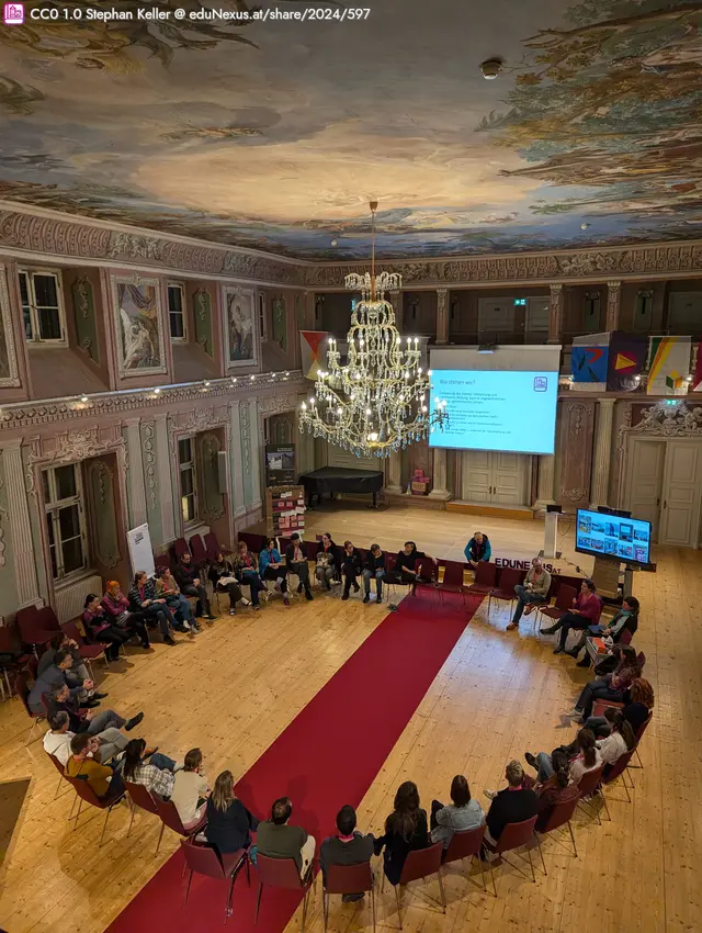 Ein großer Raum mit einem runden Teppich in der Mitte, umgeben von Stühlen mit Personen die darauf sitzen. Eine Decke mit Fresken und ein großer Kronleuchter sind sichtbar. An einer Wand steht ein Bildschirm mit Präsentationstext.