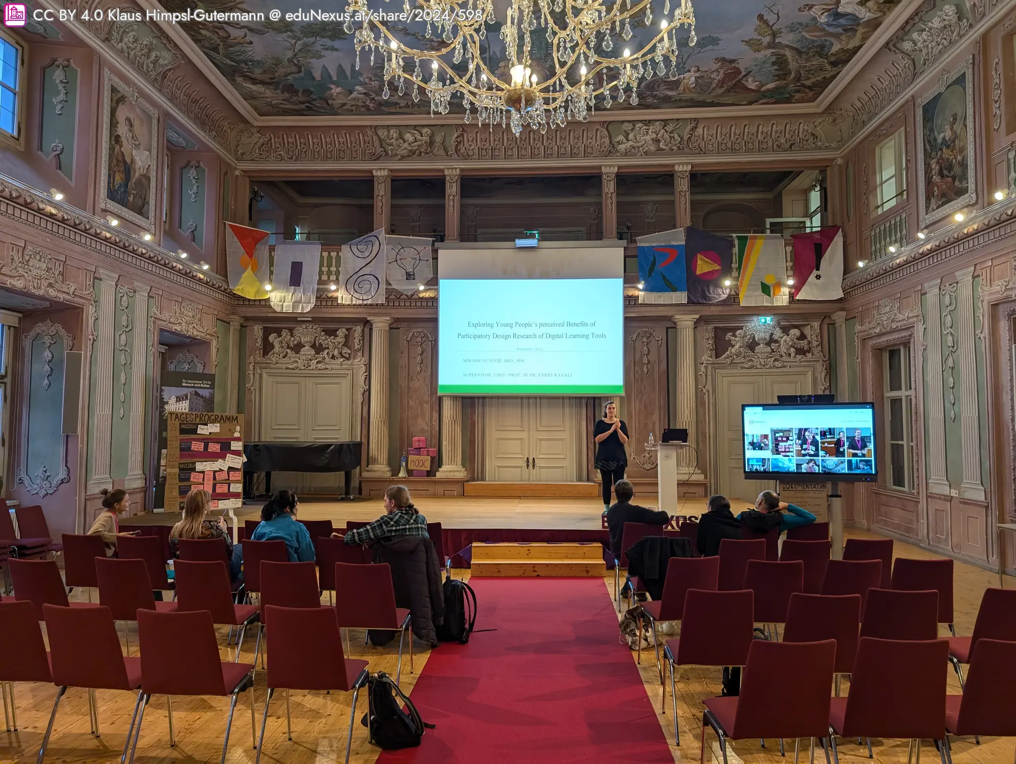 Ein Raum mit einem Kronleuchter, einer Präsentation auf einer Leinwand und mehreren Stühlen in Reihen. Eine Person steht am Podium, während das Publikum zuschaut. Bunte Fahnen hängen an der Wand.