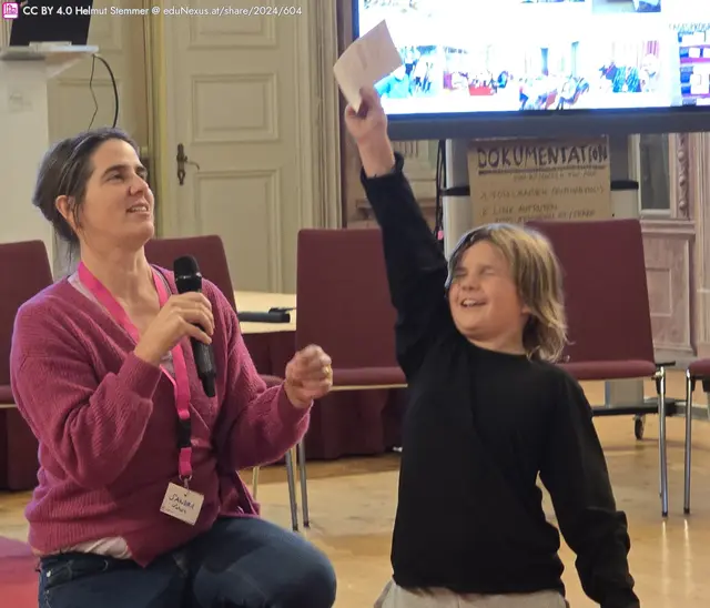 Eine Frau mit Mikrofon sitzt auf einem Stuhl und spricht, während ein Kind neben ihr mit erhobener Hand eine Karte hält und lächelt. Im Hintergrund sind Stühle und ein Bildschirm sichtbar.