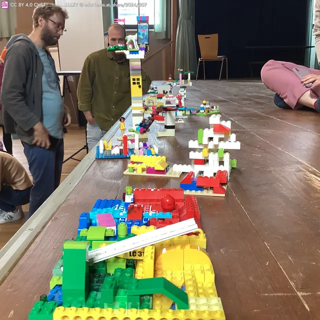 Bunte LEGO-Bauten auf einem Tisch, mit drei Personen im Hintergrund, die die Modelle betrachten.