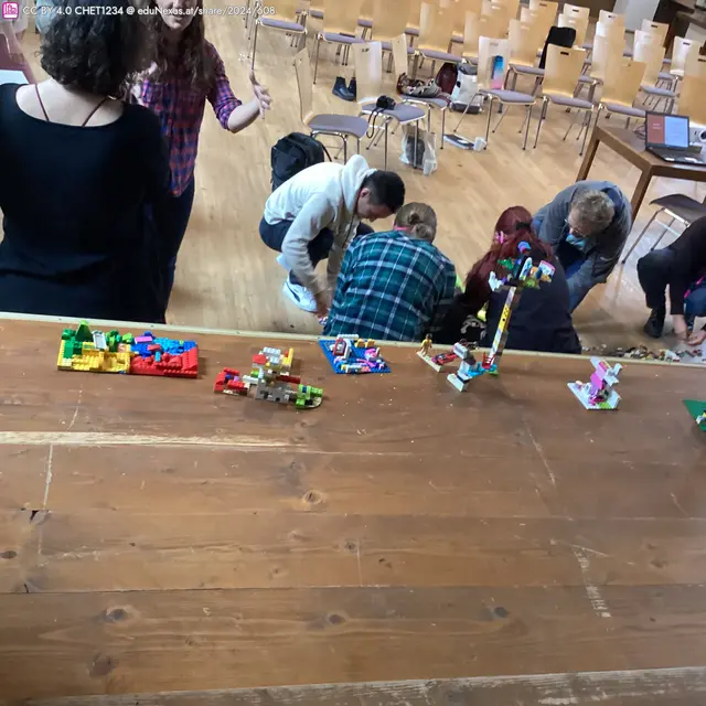 Gruppendiskussion mit mehreren Personen, die auf dem Boden sitzen und mit LEGO spielen. Im Vordergrund stehen farbige LEGO-Kreationen auf einem Tisch.